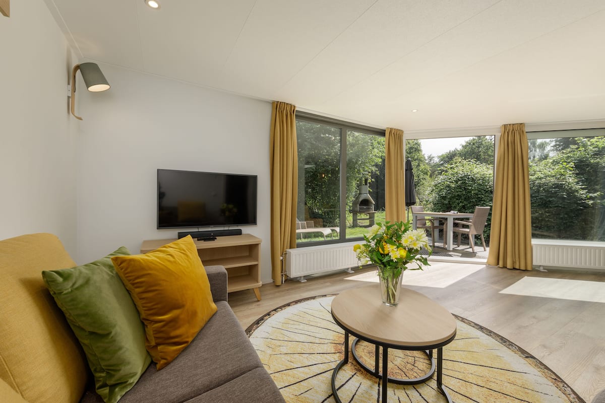 A modern living room with a flat-screen TV, a sofa with cushions, a coffee table with a vase of flowers, and a view of the outdoors through large windows.