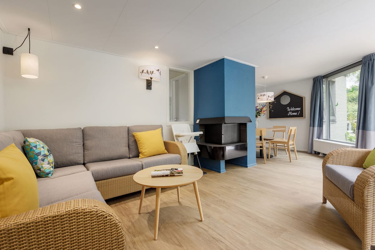 A modern living room with a sofa, coffee table, and wicker chairs. There is a fireplace, a dining table with chairs, and a sign that says "Welcome Home!".