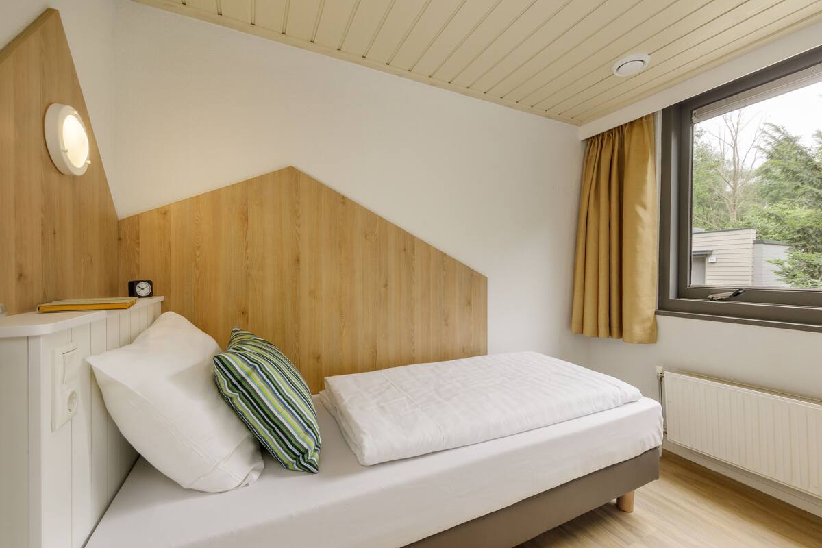 A bedroom with a wooden headboard, a bed with white bedding, a bedside table with a clock, and a window with curtains.