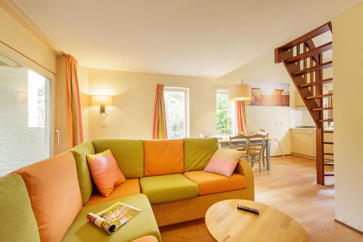 Una sala de estar acogedora con un sofá verde y naranja, una mesa de centro de madera y una cocina al fondo.