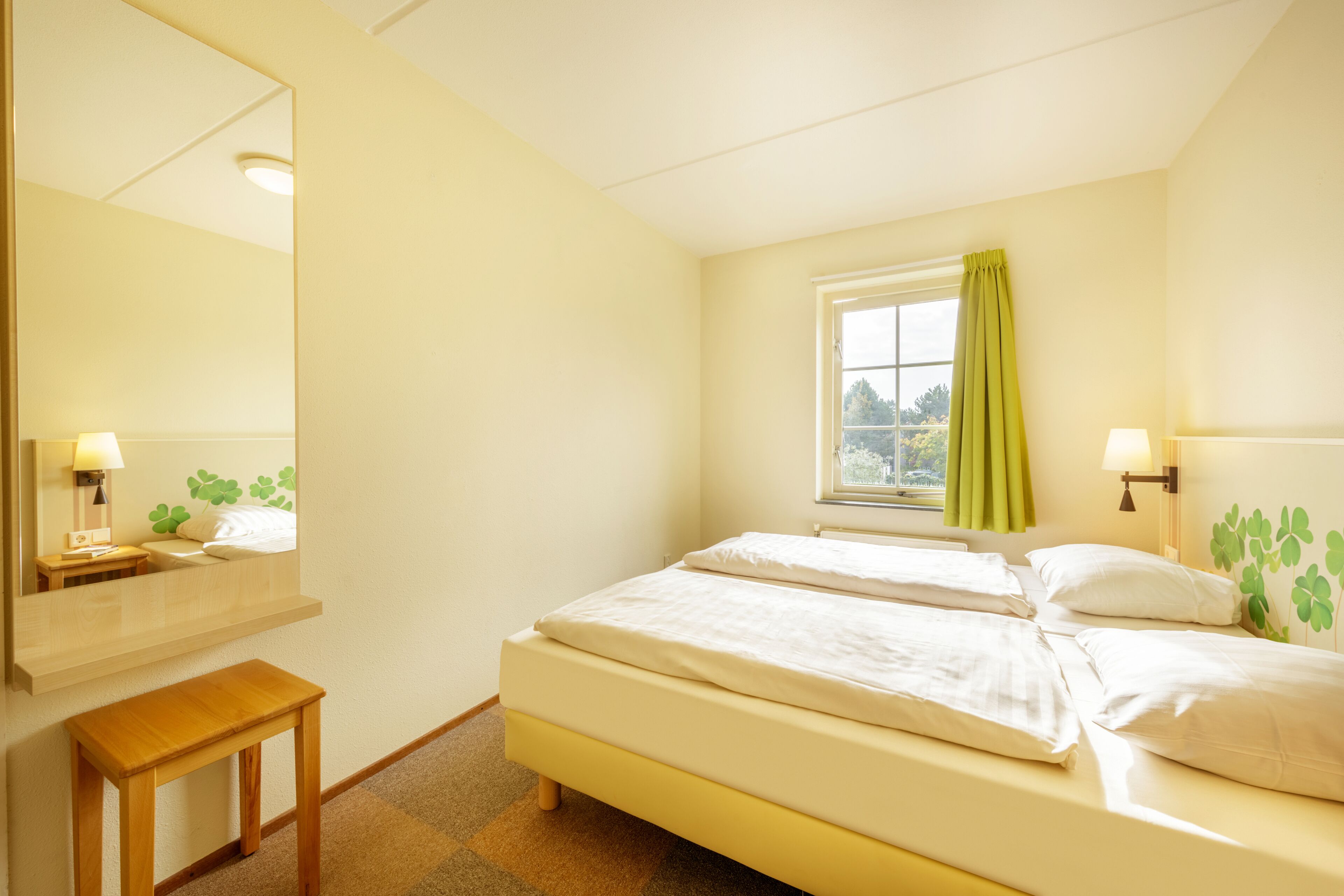 A bedroom with two beds, a window with curtains, a bedside table, and a mirror.