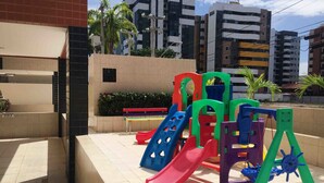 Un parque infantil con toboganes y columpios, rodeado de edificios y una zona de patio con baldosas.