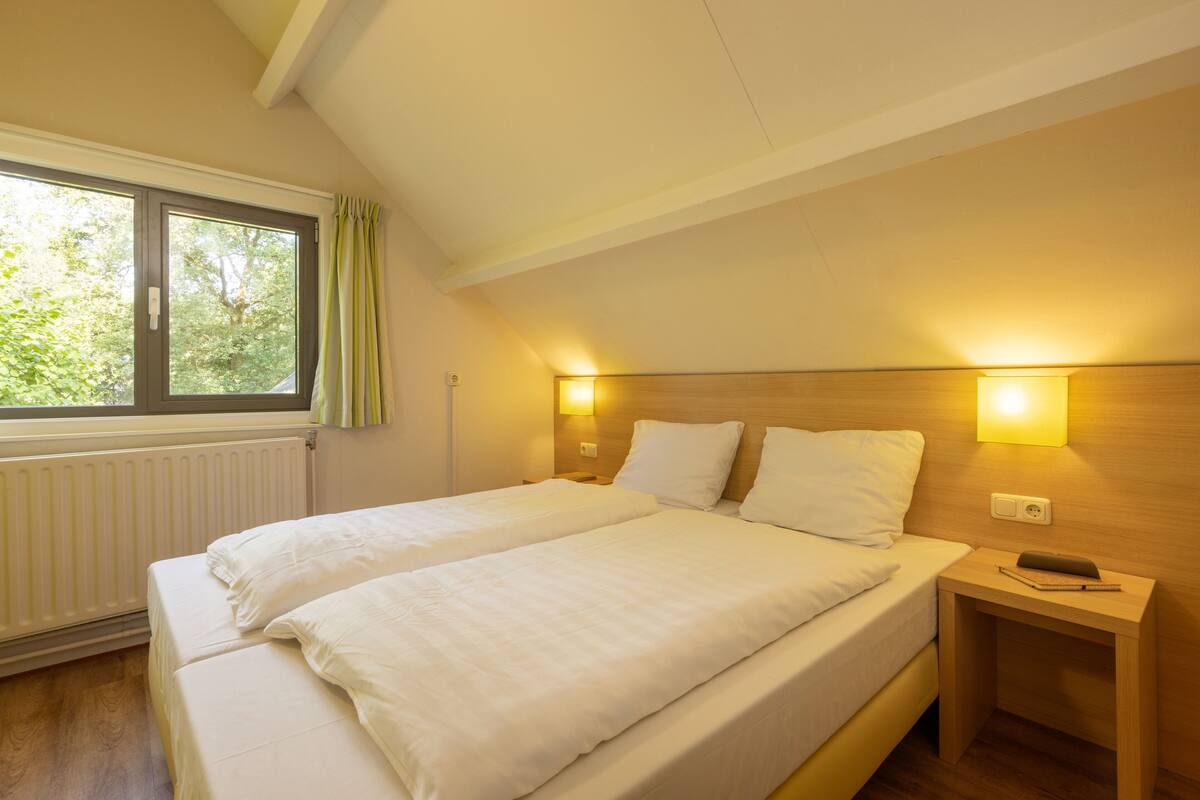 A double bed with white linens, a bedside table with a lamp, and a window with curtains.