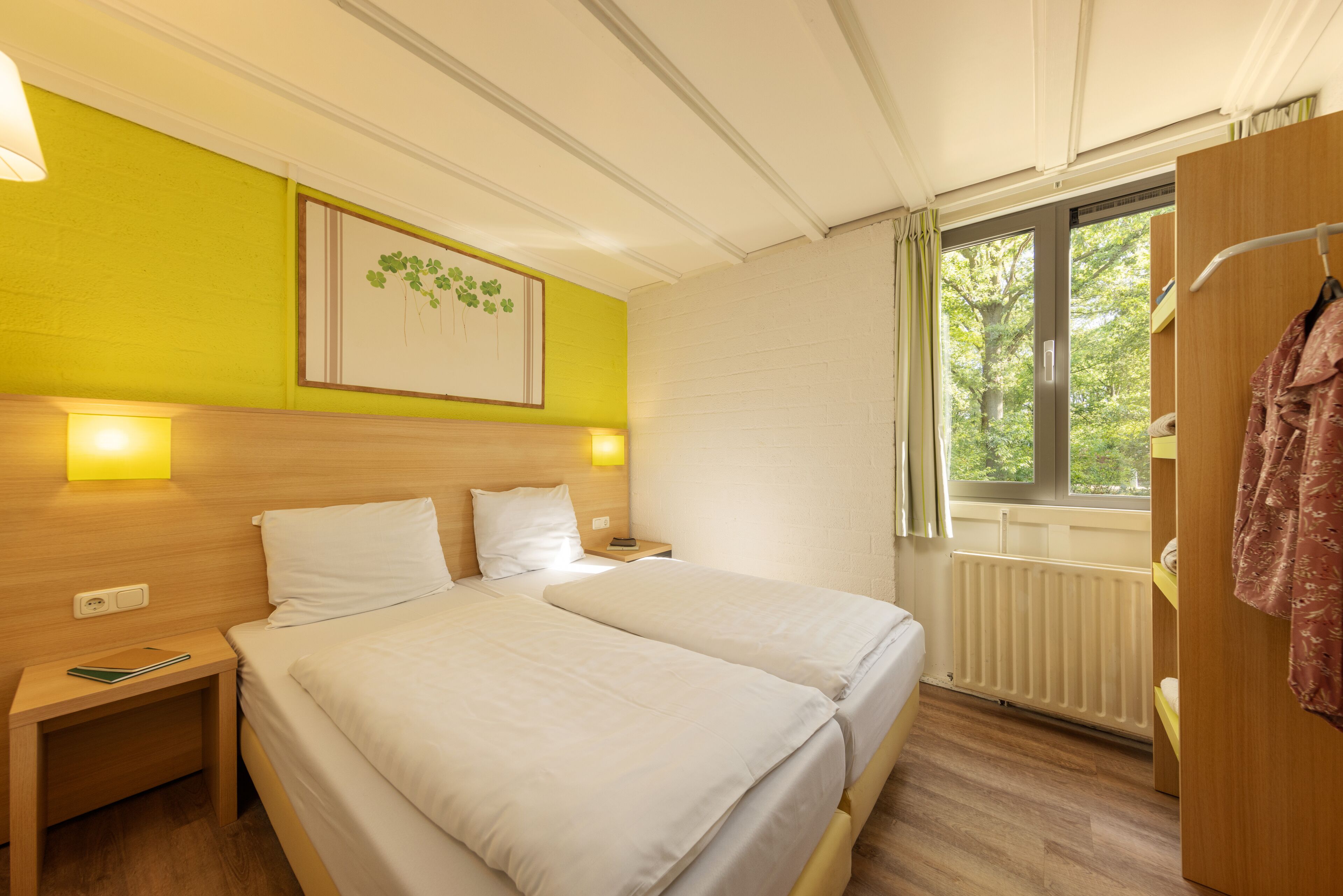 A hotel room with two beds, a window with curtains, a radiator, and a wall-mounted picture.