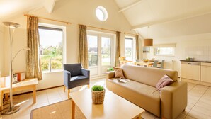 A spacious living area with a sofa, armchair, and a dining table. There is a kitchen area with cabinets and a window overlooking a garden. - Center Parcs Parc Sandur (Emmen)