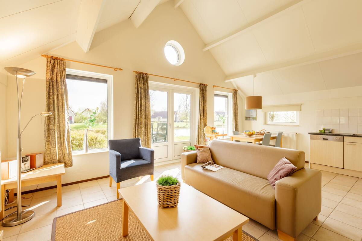 A spacious living area with a sofa, armchair, and a dining table. There is a kitchen area with cabinets and a window overlooking a garden.