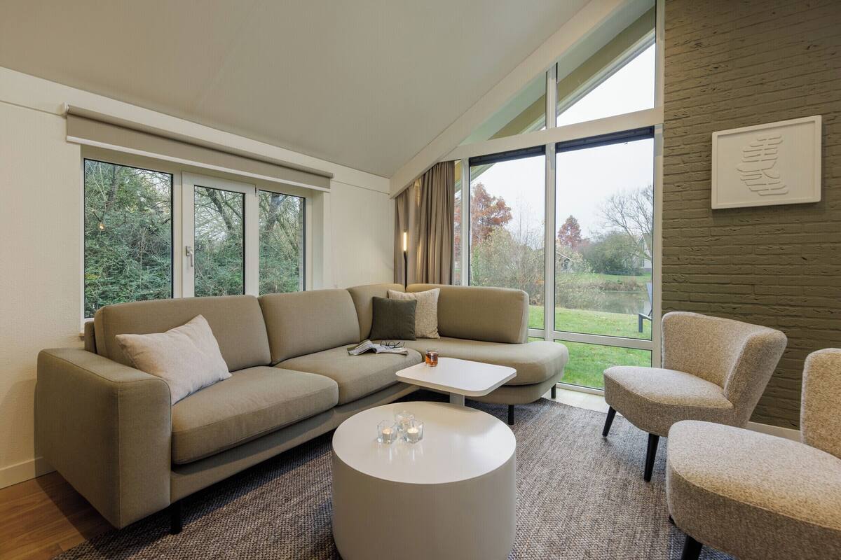 A modern living room with a sectional sofa, a round coffee table, and two armchairs. Large windows provide a view of the outdoors.