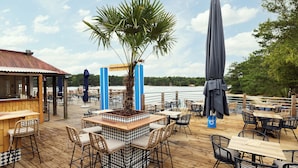 An outdoor dining area with wooden tables and chairs, a central planter with a palm tree, and umbrellas providing shade. - Center Parcs De Vossemeren (Lommel)