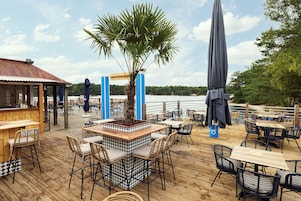 An outdoor dining area with wooden tables and chairs, a central planter with a palm tree, and umbrellas providing shade.