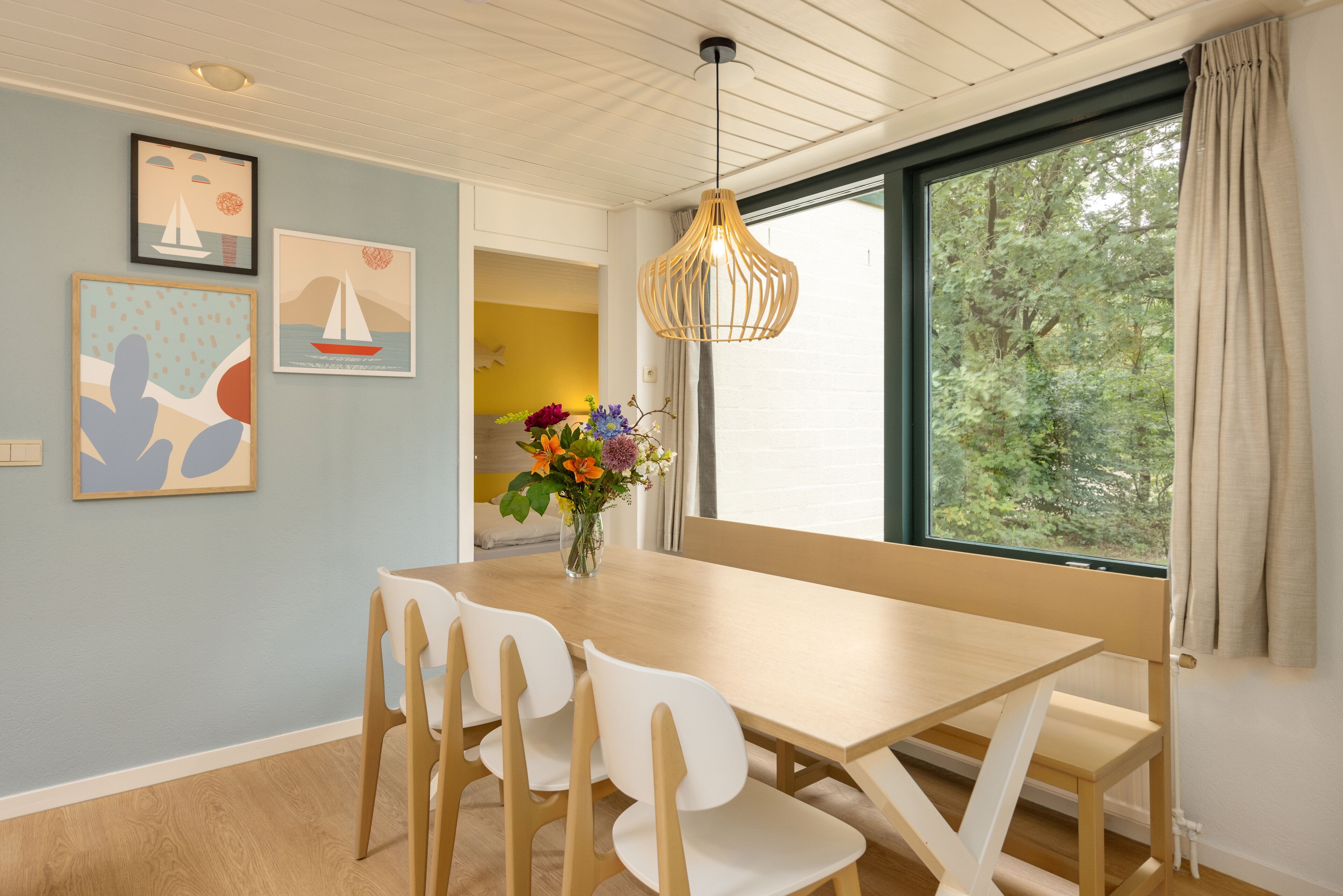 Een eetgedeelte met een houten tafel en witte stoelen, een hanglamp en kunstwerken in lijsten aan de wand.