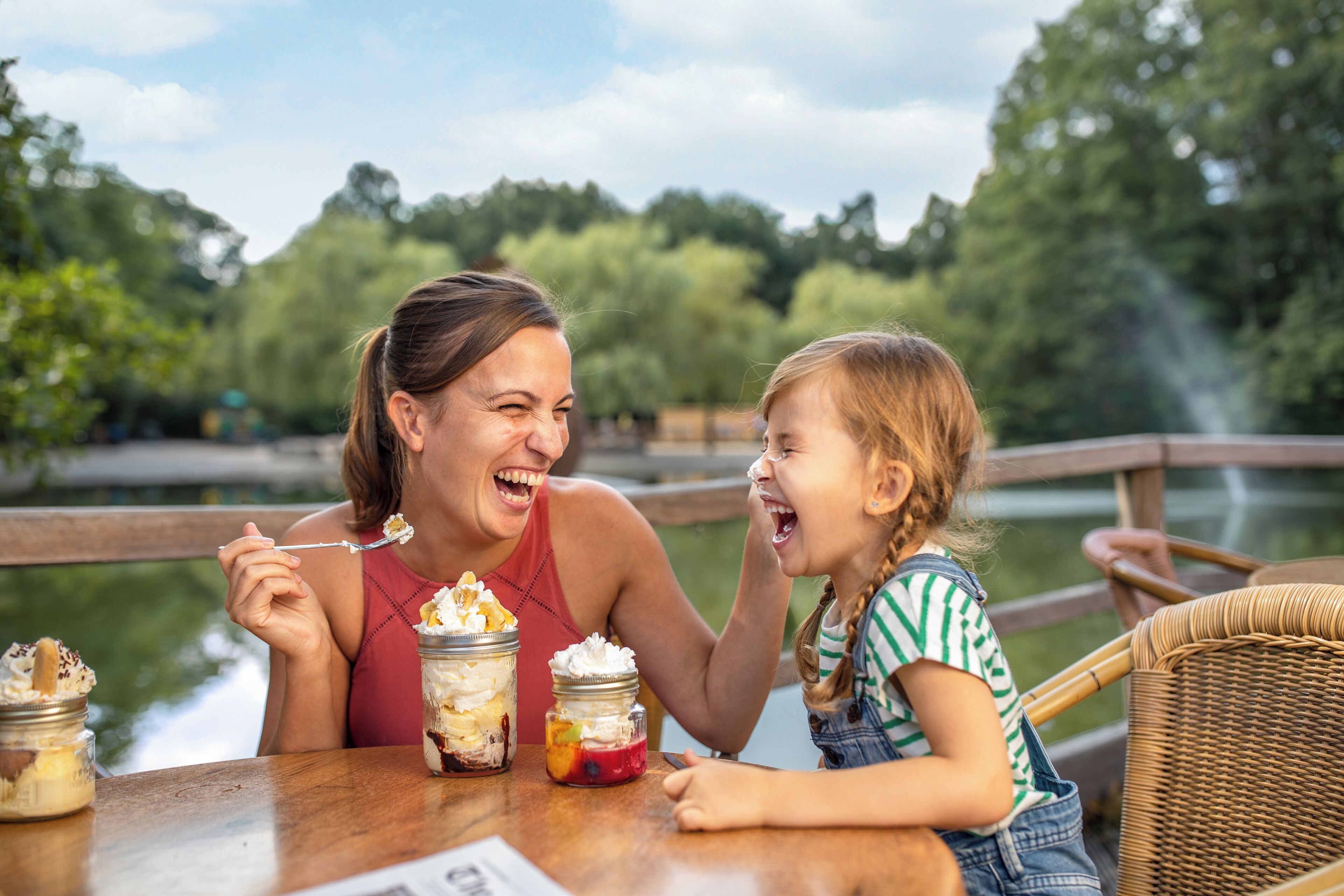 Twee personen zitten aan een houten tafel met desserts voor zich, buiten met een houten leuning en groen op de achtergrond.