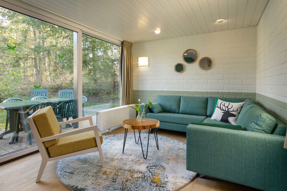 A cozy living room with a green sofa, a wooden coffee table, and a chair. There is a large window providing a view of the outdoors.
