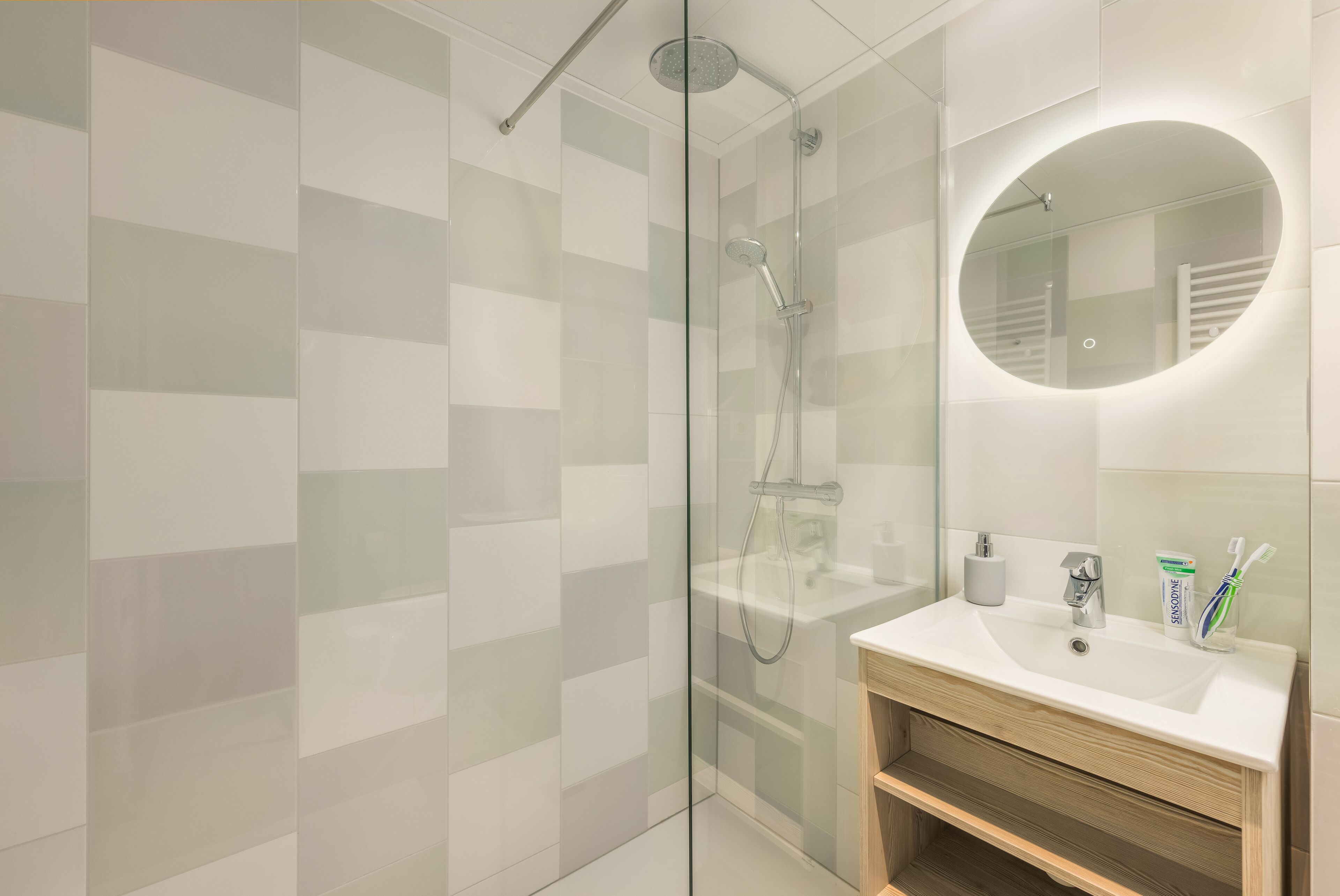 Een moderne badkamer met een douchecabine van glas, een ronde spiegel en een houten wastafelmeubel met een wastafel.