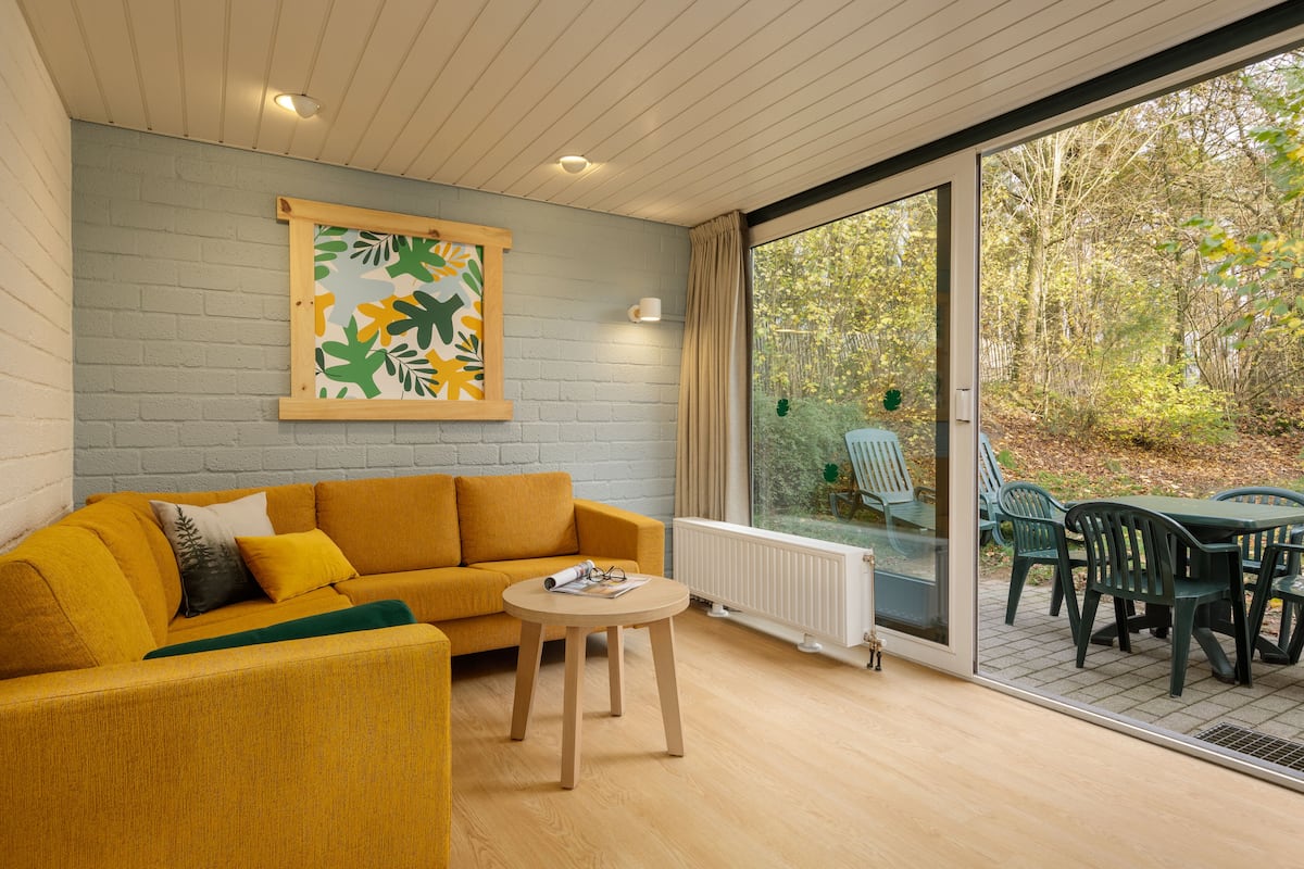 A modern living room with a yellow sofa, a round wooden table, and a large window overlooking a garden.