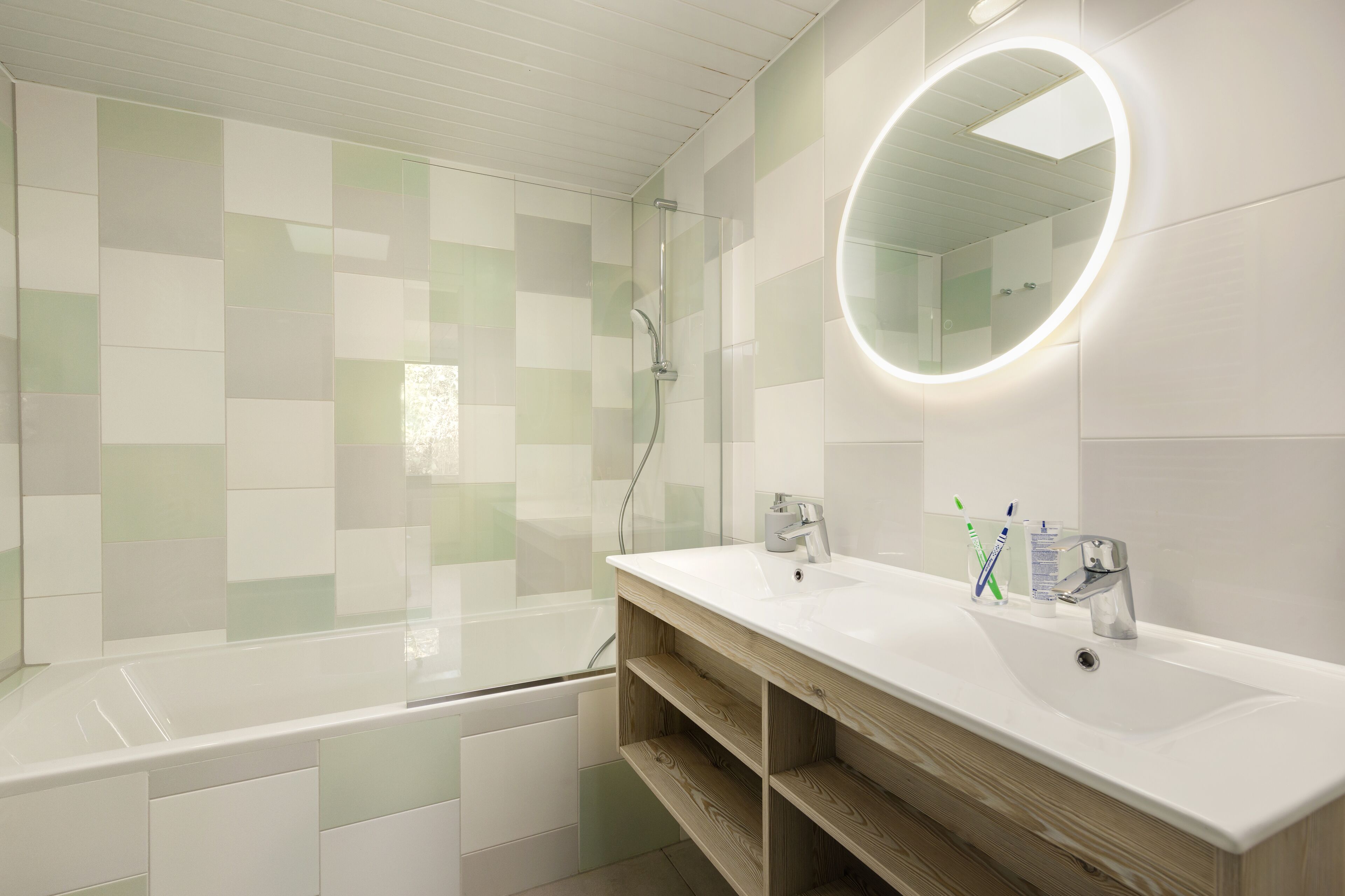 Een moderne badkamer met een glazen douchecabine, een witte wastafel met houten opbergruimte en een ronde spiegel met verlichting.