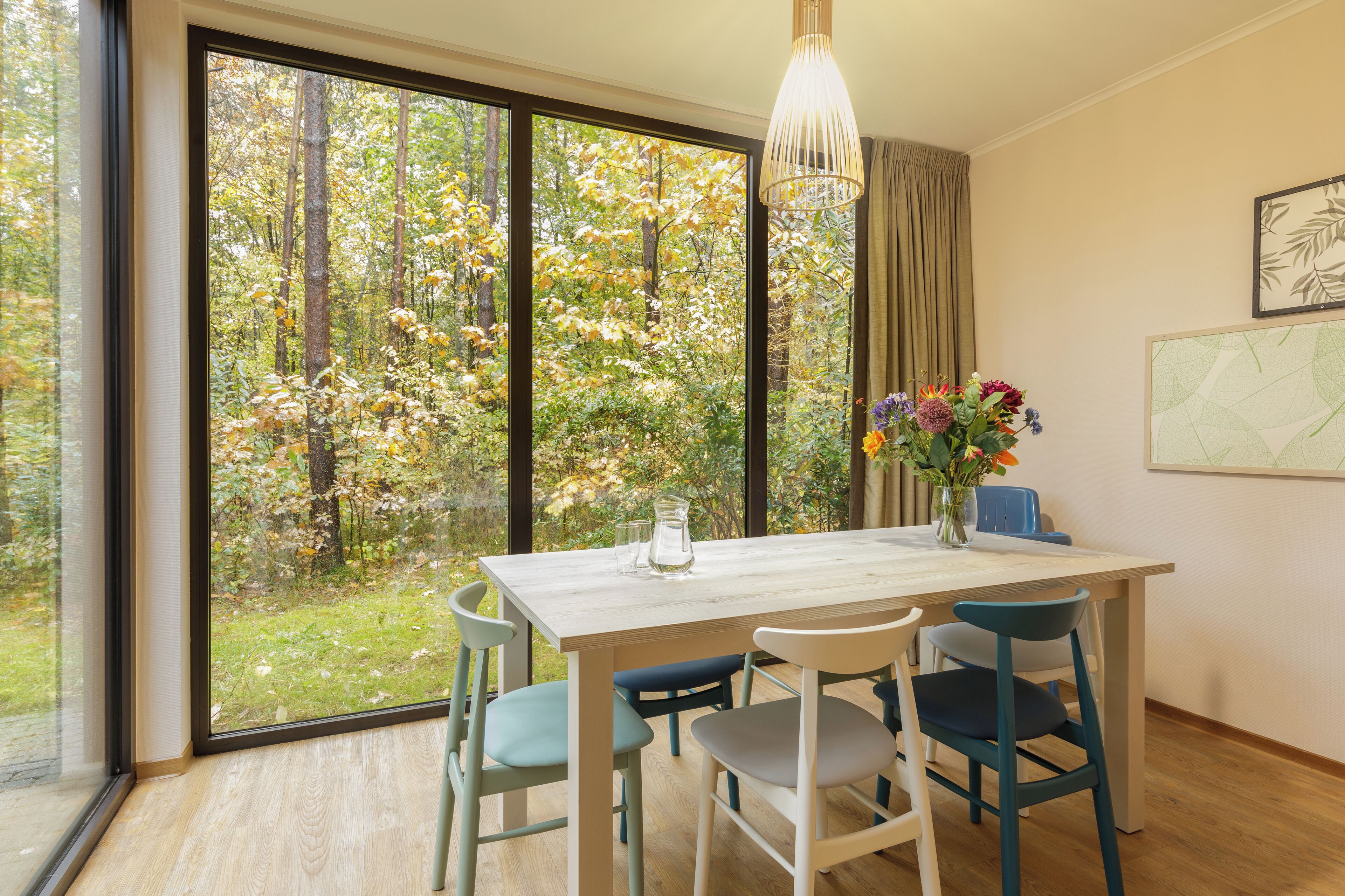 A dining area with a table, chairs, a vase of flowers, and a view of a forest through large windows.