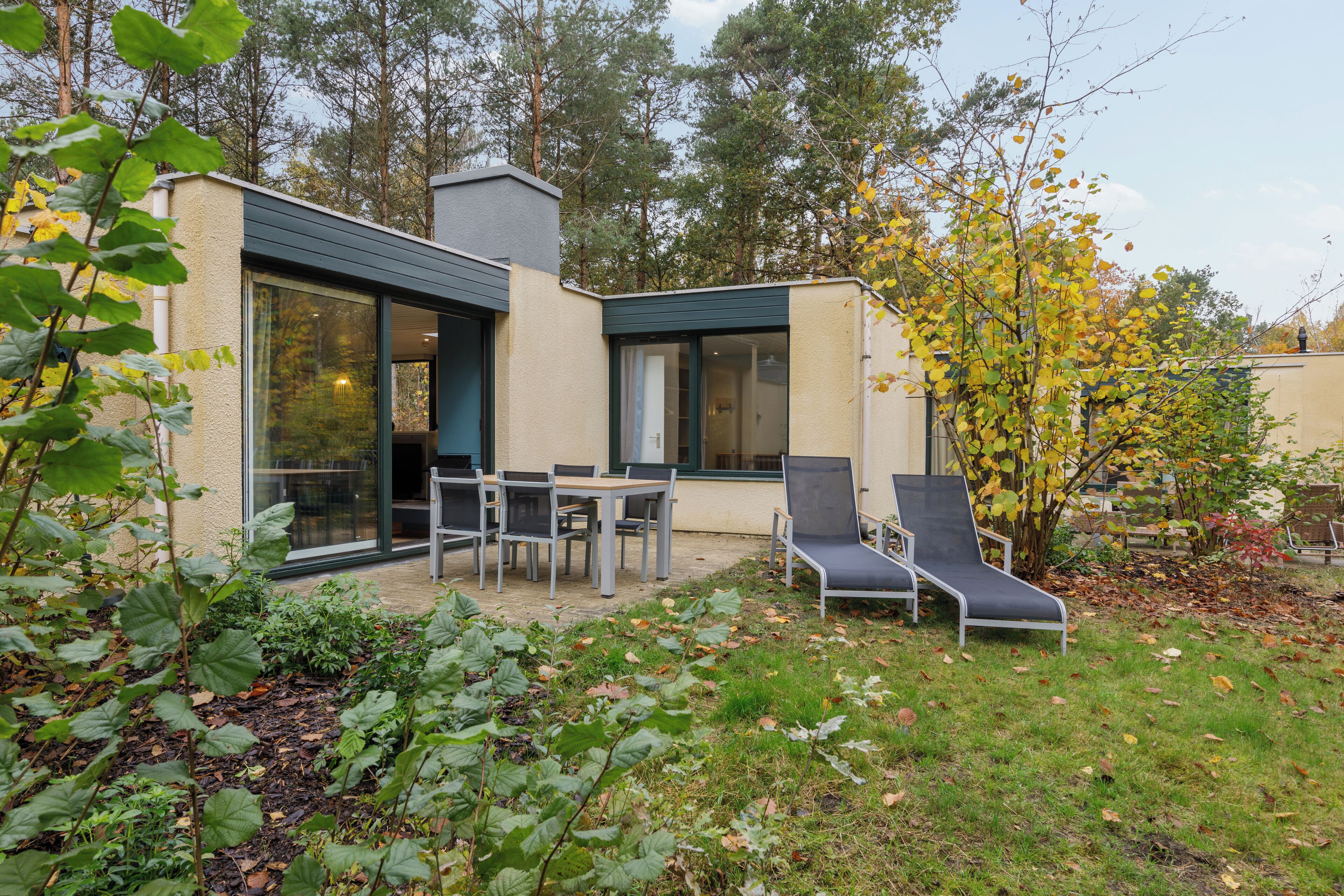 Ein modernes Haus mit einer Terrassenanlage, inklusive Tisch, Stühlen und einer Liege, sowie ein Garten mit Pflanzen und Bäumen.
