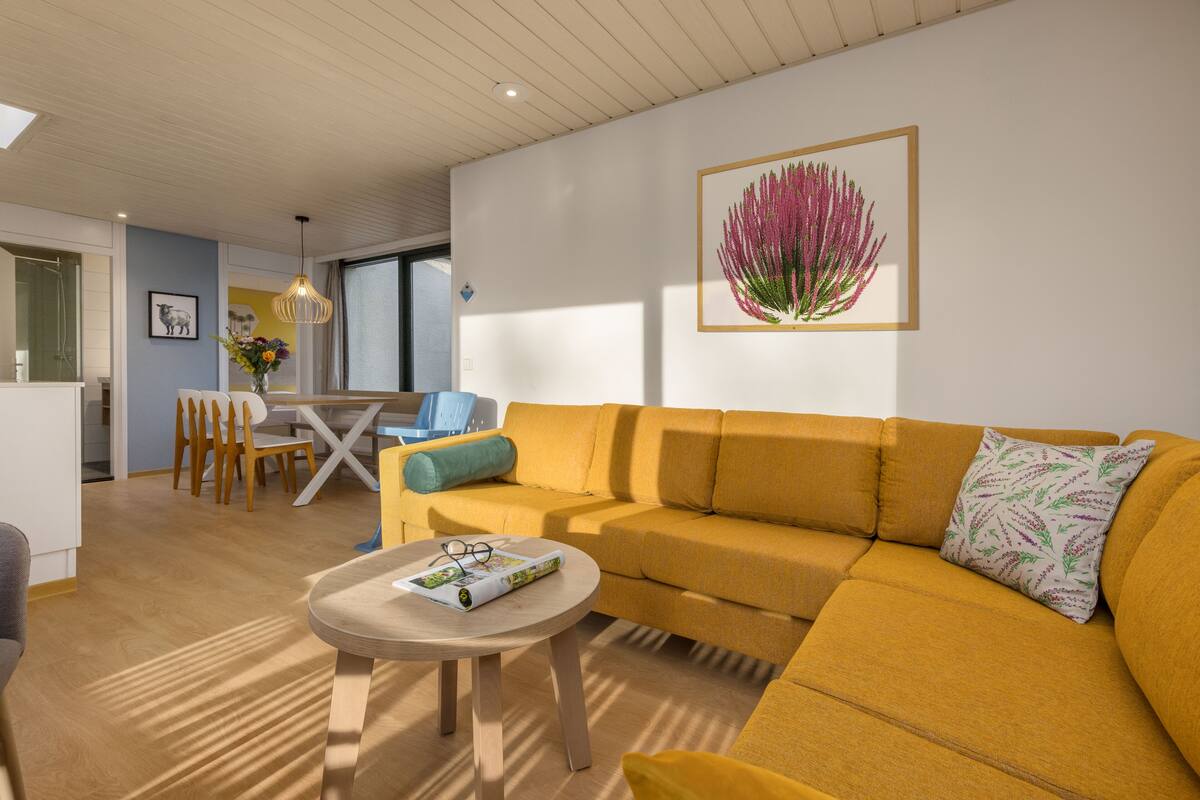 En moderne stue med en gul sofa, et rundt træbord og et spiseområde med et hvidt bord og stole.