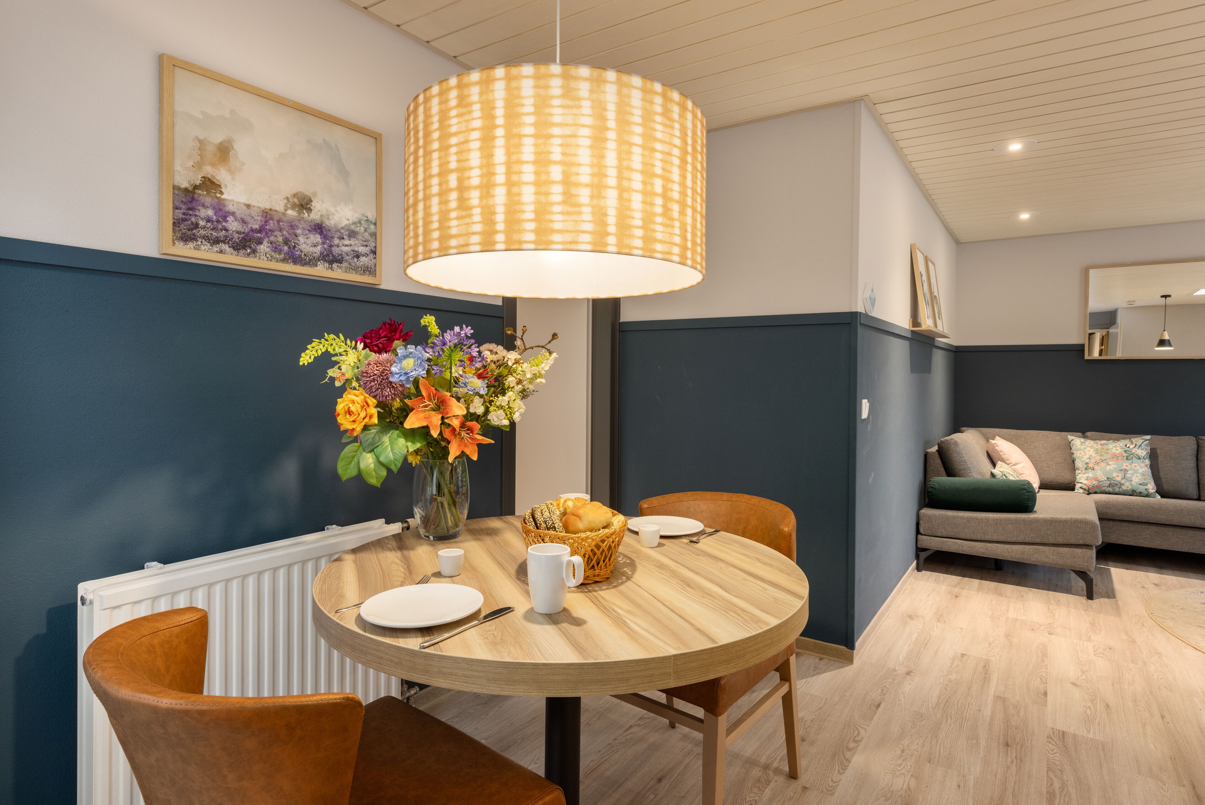 Een eetgedeelte met een ronde tafel, stoelen, een bloemenvaas en een hanglamp.