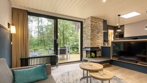 Un salon moderne comprenant un canapé, une table ronde, une cheminée, un téléviseur et une grande fenêtre donnant sur des arbres.
