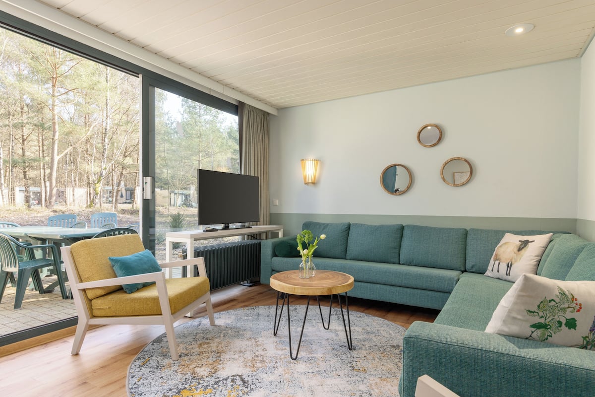 En moderne stue med en grøn sofa, et træbord og en gul lænestol. Der er et stort vindue med udsigt til træer og et spiseområde med stole og et bord.