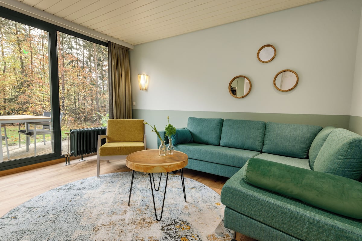 En moderne stue med en grøn sofa, en gul lænestol og et rundt træbord. Der er tre vægmonterede spejle og et stort vindue med udsigt til træer.