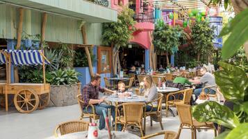 Relax in a casual atmosphere with wicker chairs and round tables amidst lush greenery and a vibrant building facade.