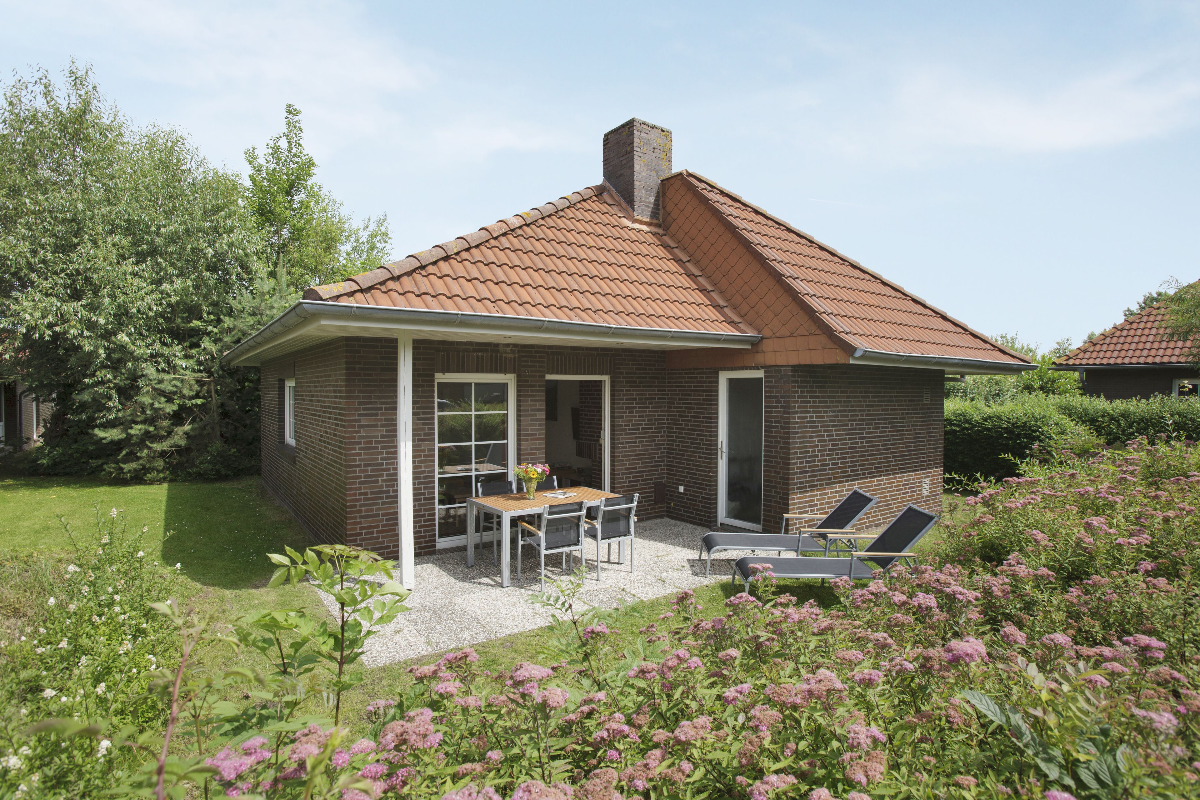 Une petite maison en briques avec un toit en tuiles, une terrasse avec une table et des chaises, et un jardin parsemé de fleurs roses.
