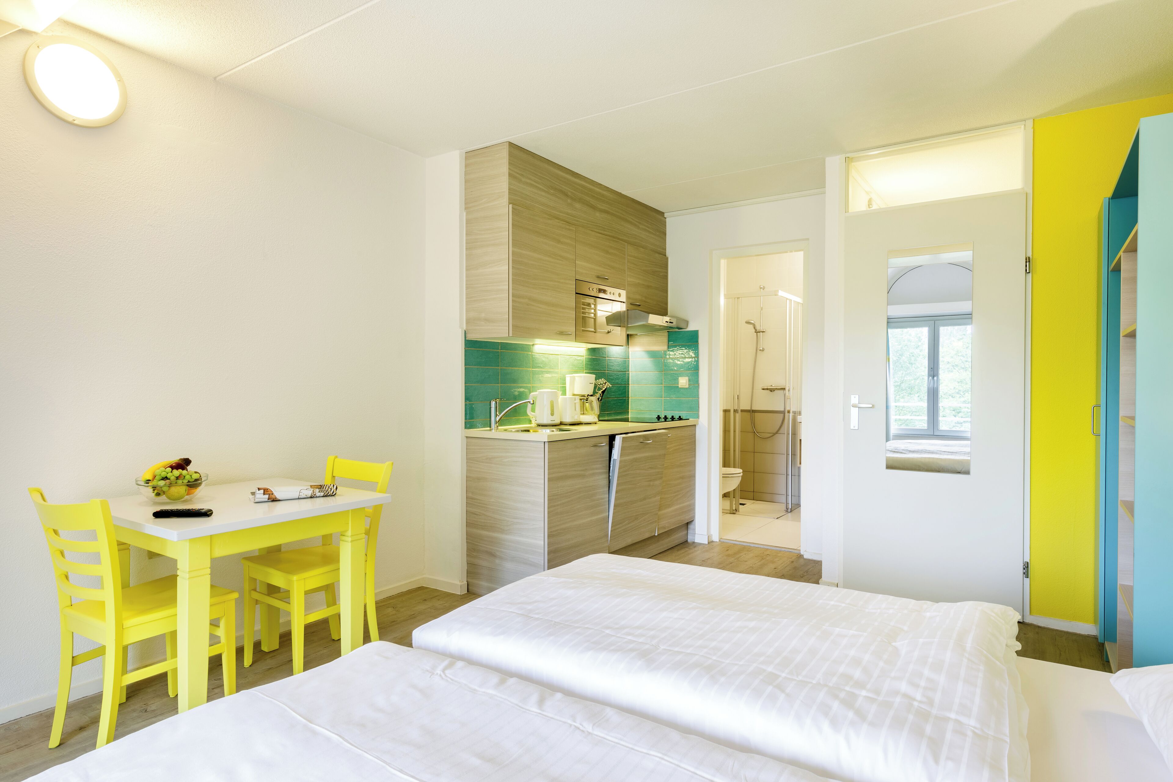 Ein Hotelzimmer mit Bett, einem kleinen Esstisch mit Stühlen, einer Küchenzeile und einem durchgehenden Türspalte sichtbaren Badezimmer.
