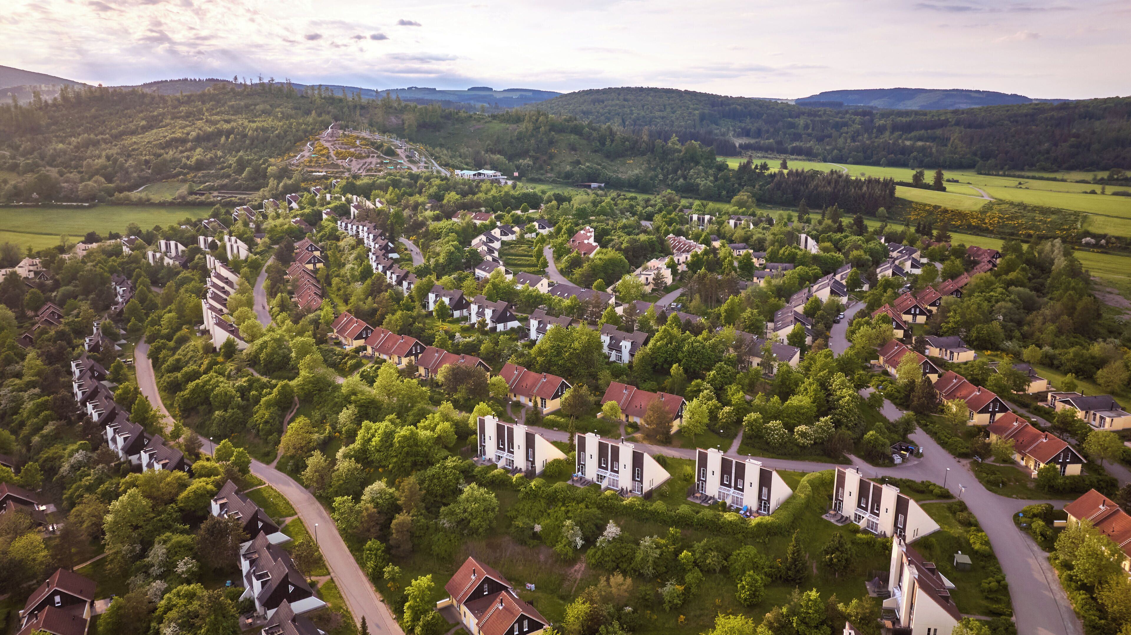 An aerial view of a residential area with houses arranged in a circular pattern, surrounded by greenery and hills in the background.
