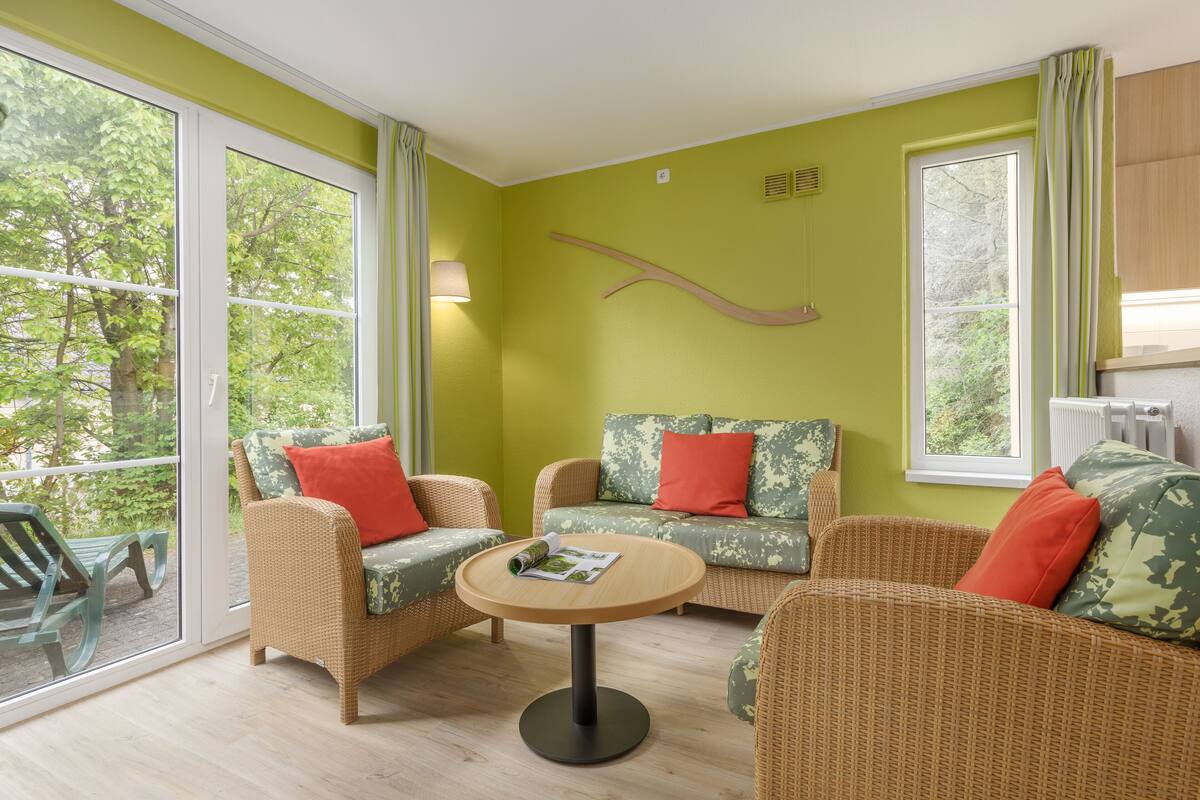 Une pièce meublée de mobilier en osier, avec une table ronde et une vue sur la verdure grâce à de grandes fenêtres.