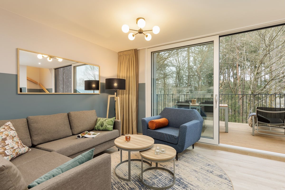 Un salon moderne comprenant un canapé, un fauteuil, deux tables rondes, un grand miroir et une porte-fenêtre coulissante donnant sur un balcon extérieur.