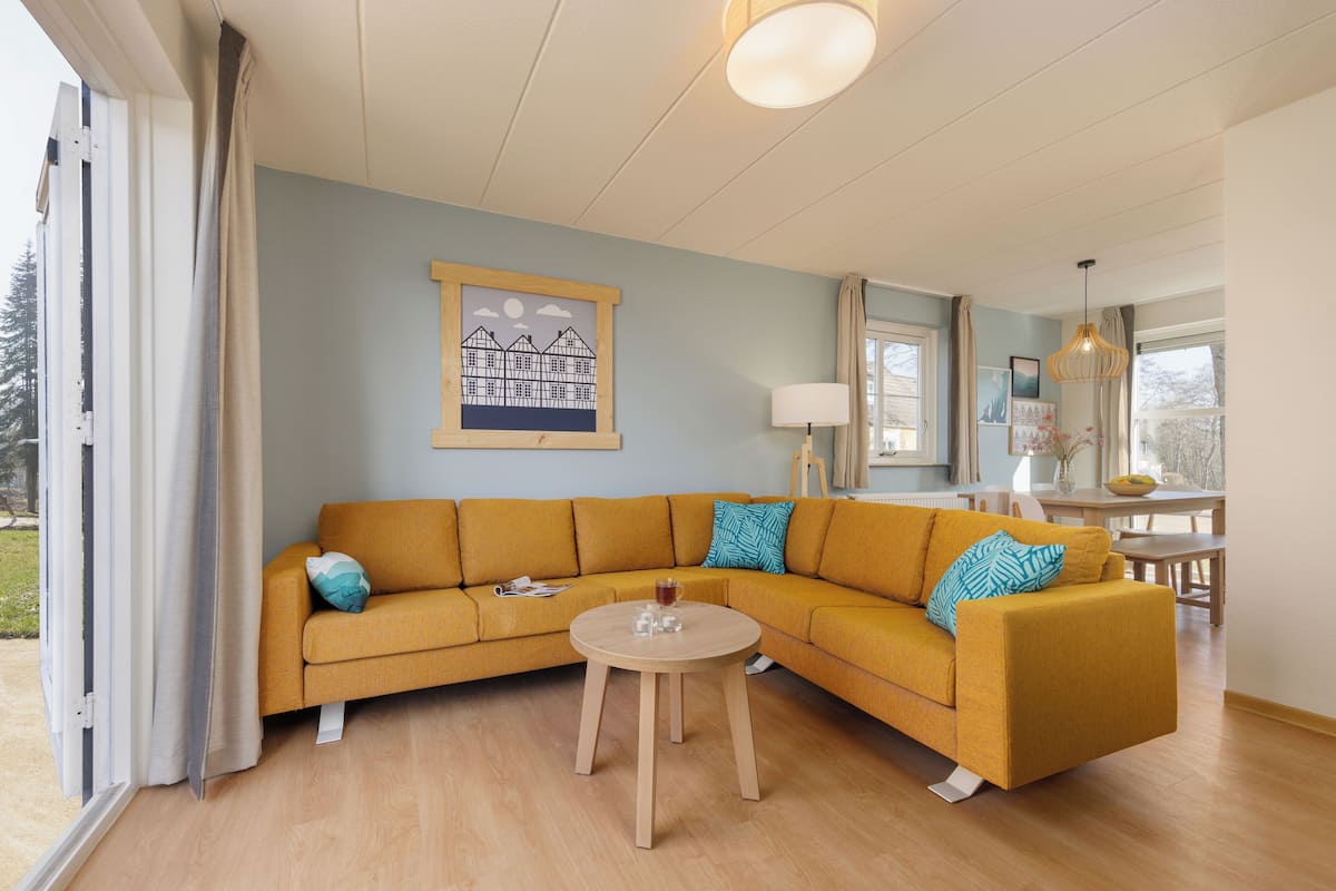 Un salon moderne avec un canapé de couleur moutarde, une table basse ronde en bois et une œuvre d’art encadrée accrochée au mur.