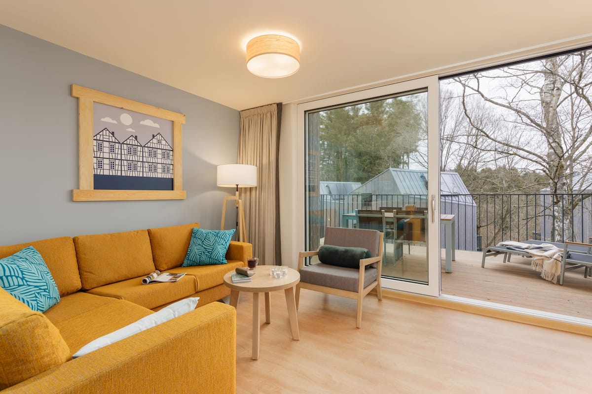 Un salon moderne avec un canapé, un fauteuil et une petite table. Une grande fenêtre offre une vue sur une terrasse et des arbres.