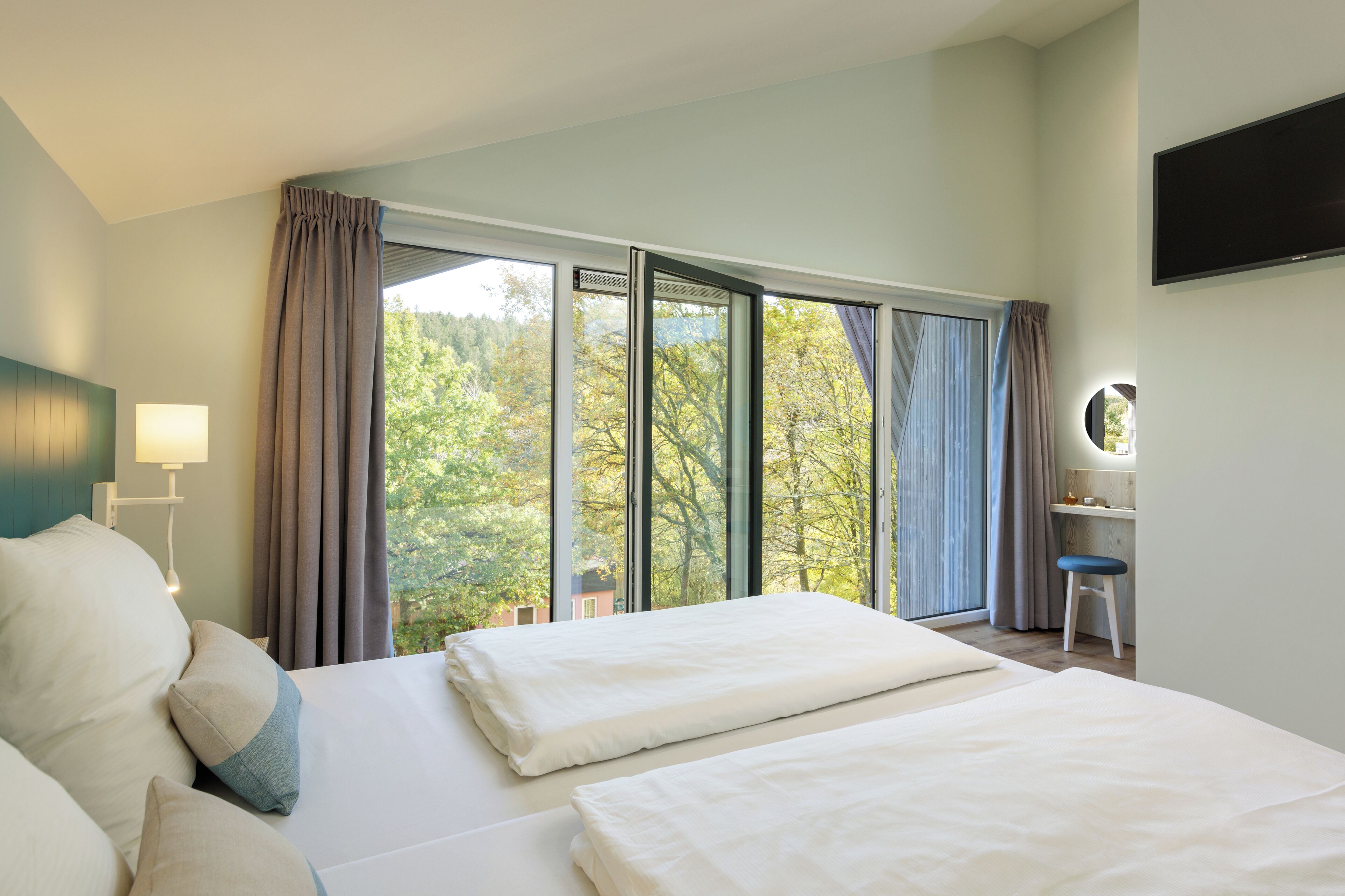 Ein Hotelzimmer mit zwei Betten, einem Schreibtisch mit Stuhl, einem Fernseher und einem großen Fenster mit Vorhängen.