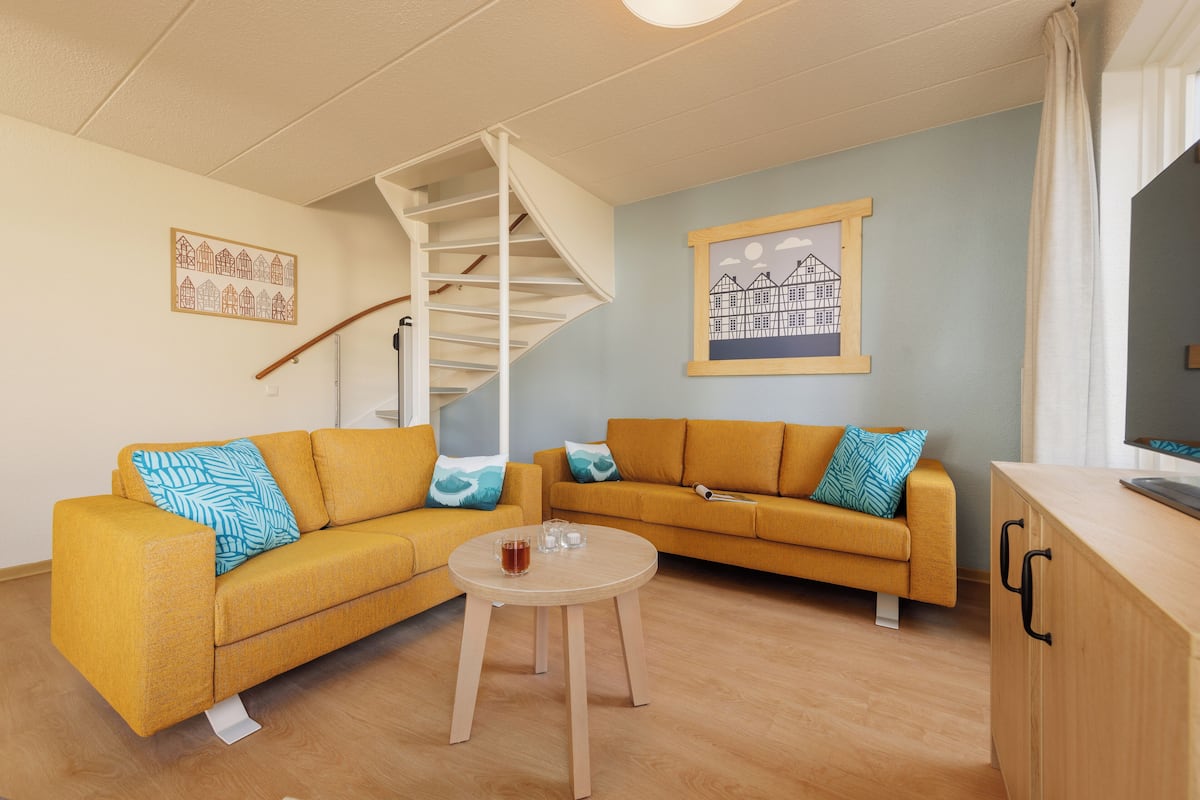 Un salon avec deux canapés de couleur moutarde, une petite table ronde, un escalier en colimaçon blanc et un tableau encadré au mur.