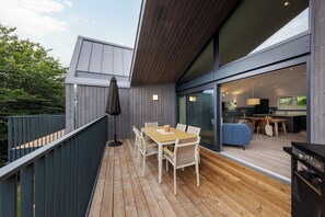 A modern outdoor deck with a wooden table and chairs, a dark umbrella, and a view into the interior of a contemporary home. - Center Parcs Park Eifel (Gunderath)