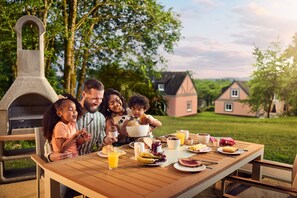 A family dining at an outdoor table with a stone oven, surrounded by greenery and houses. - Center Parcs Park Eifel (Gunderath)