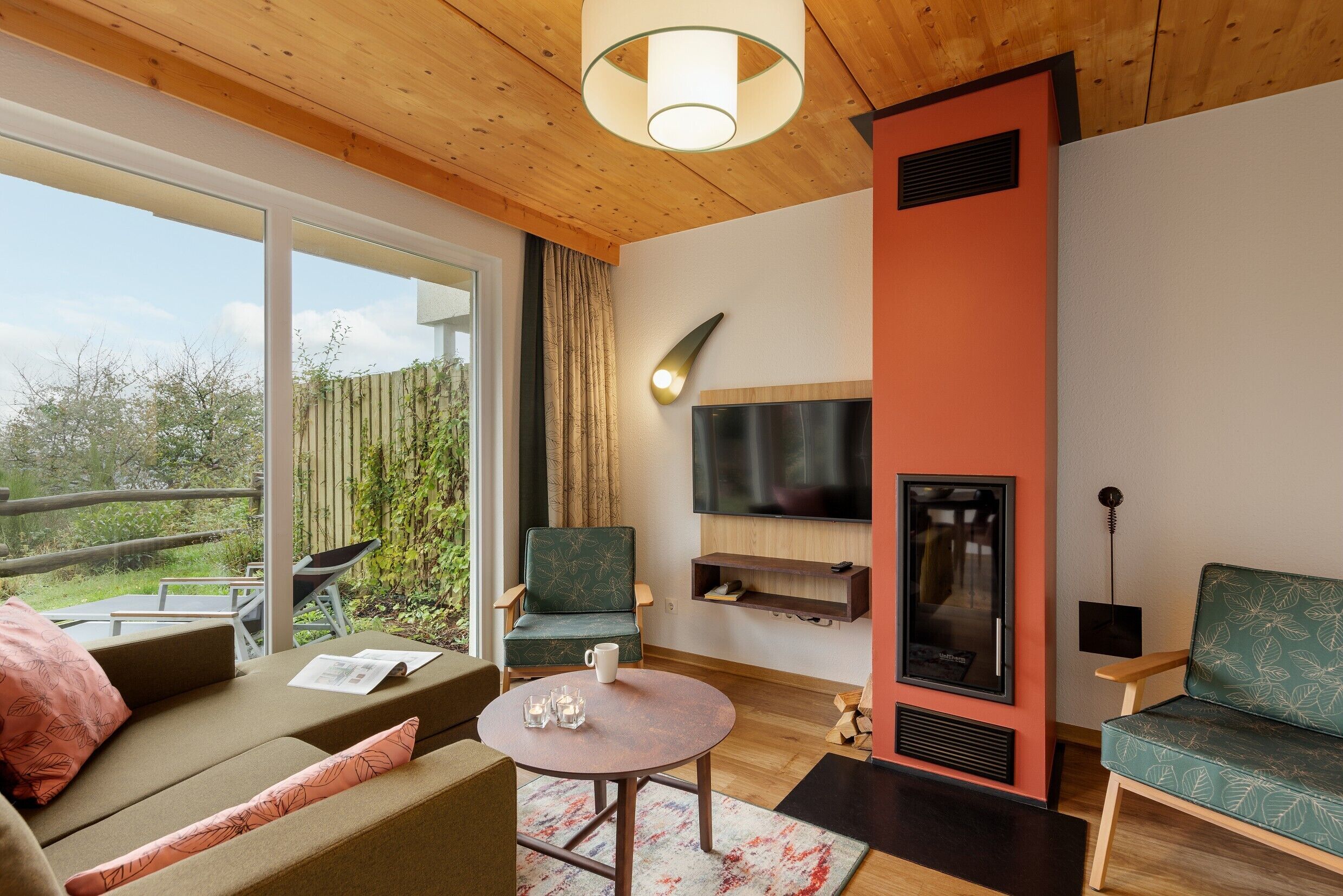 Ein modernes Wohnzimmer mit einer Couch, einem runden Couchtisch und einem an der Wand montierten Fernseher. Ein Kamin ist zu sehen, und durch die Glastür gelangt man auf einen Balkon.