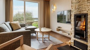 En hyggelig stue med pejs, sofa, sofabord og et stort vindue med udsigt til naturen.