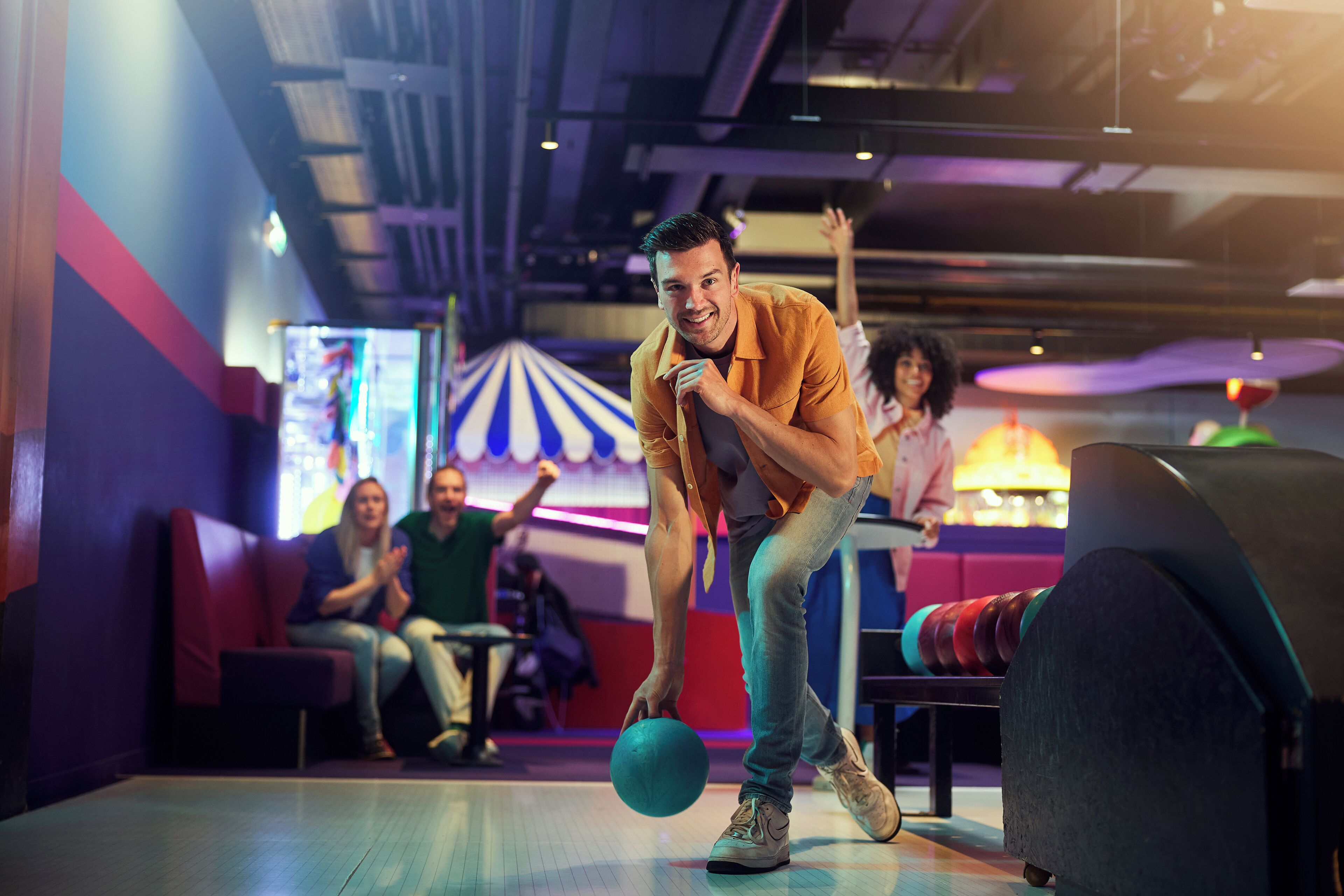 Un bowling où une personne portant un maillot orange se prépare à lancer la boule. On aperçoit des zones où sont assis des spectateurs.