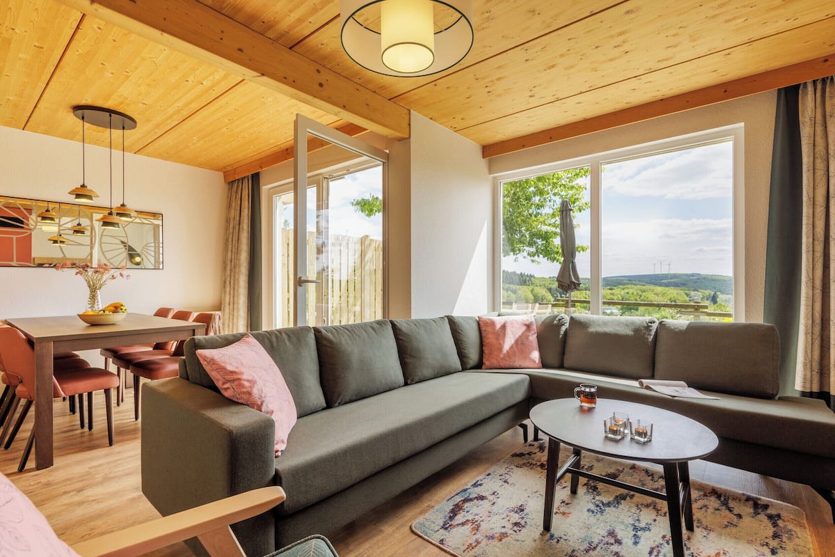 A modern living room with a sectional sofa, a dining table with chairs, and large windows offering a view of the outdoors.