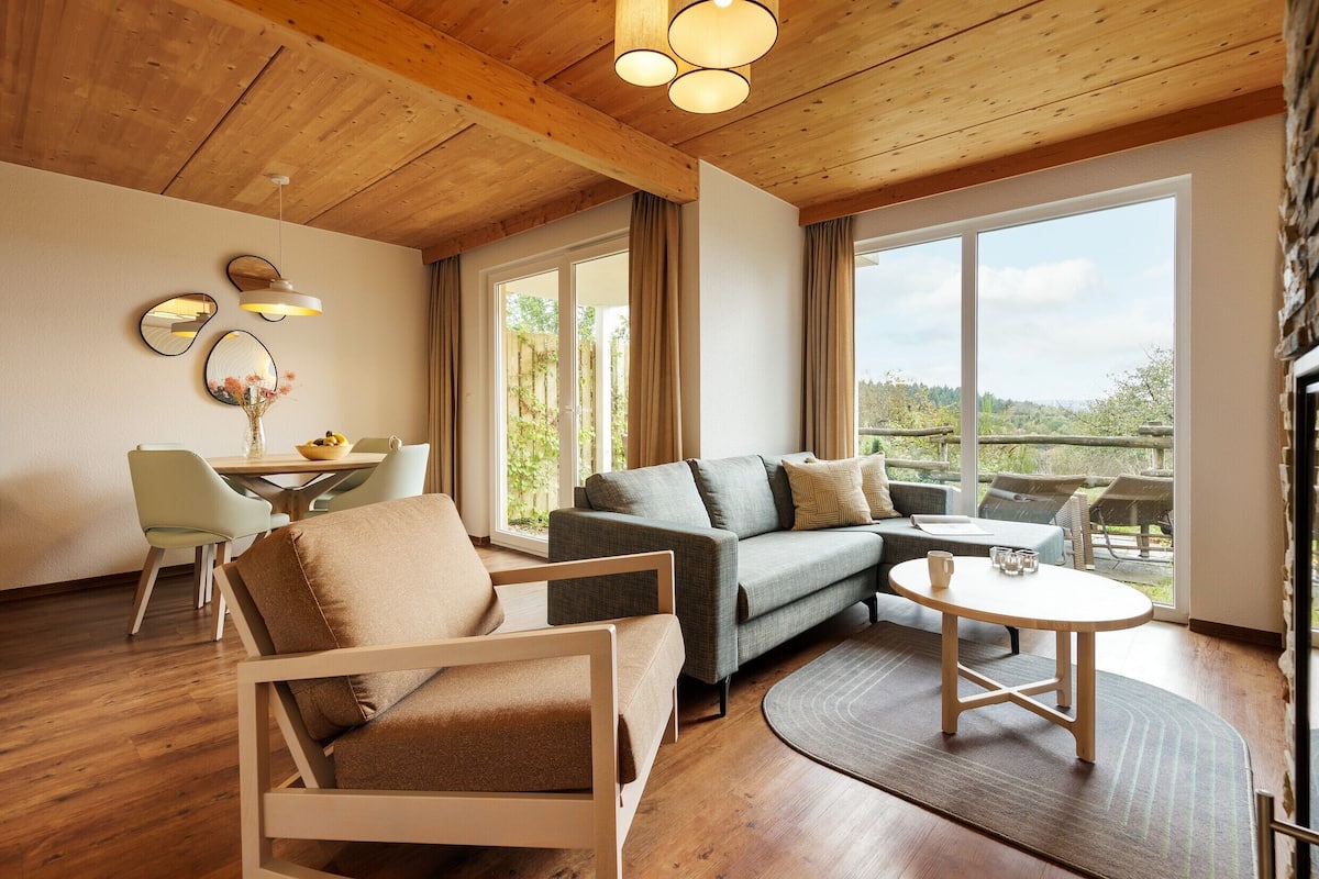 A modern living room with a sofa, armchair, and round table, wooden flooring, and large windows offering a view of the outdoors.