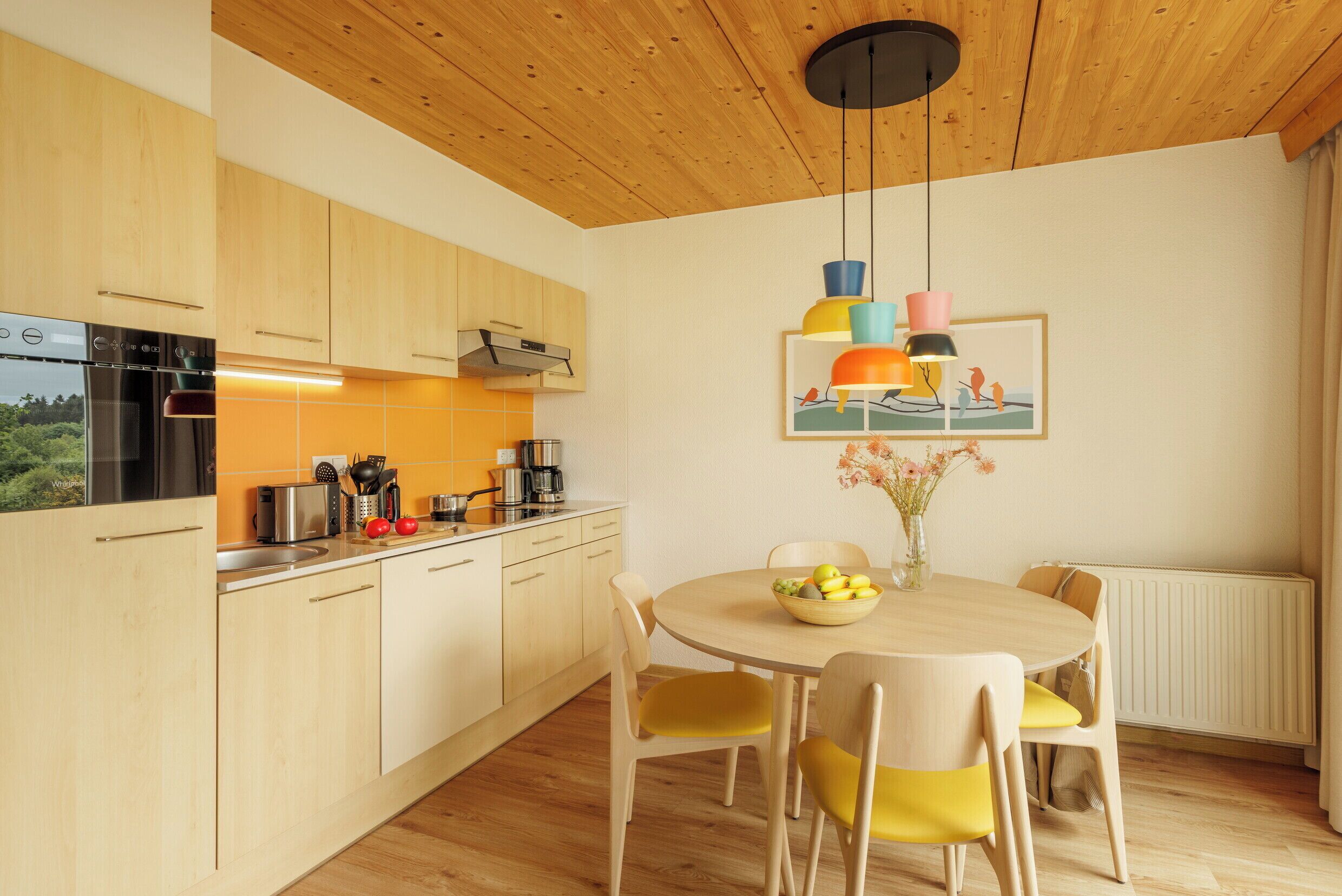 Une cuisine moderne avec des meubles en bois, un four encastré et un coin repas comprenant une table ronde et des chaises jaunes.