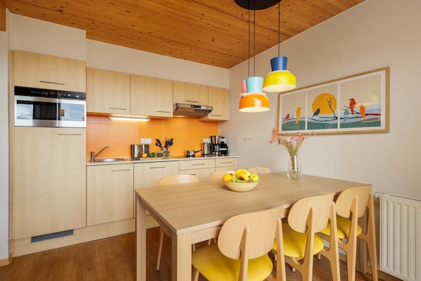 A modern kitchen with wooden cabinets, a built-in oven, and a dining area with a wooden table and yellow chairs. - Center Parcs Park Bostalsee (Nohfelden)