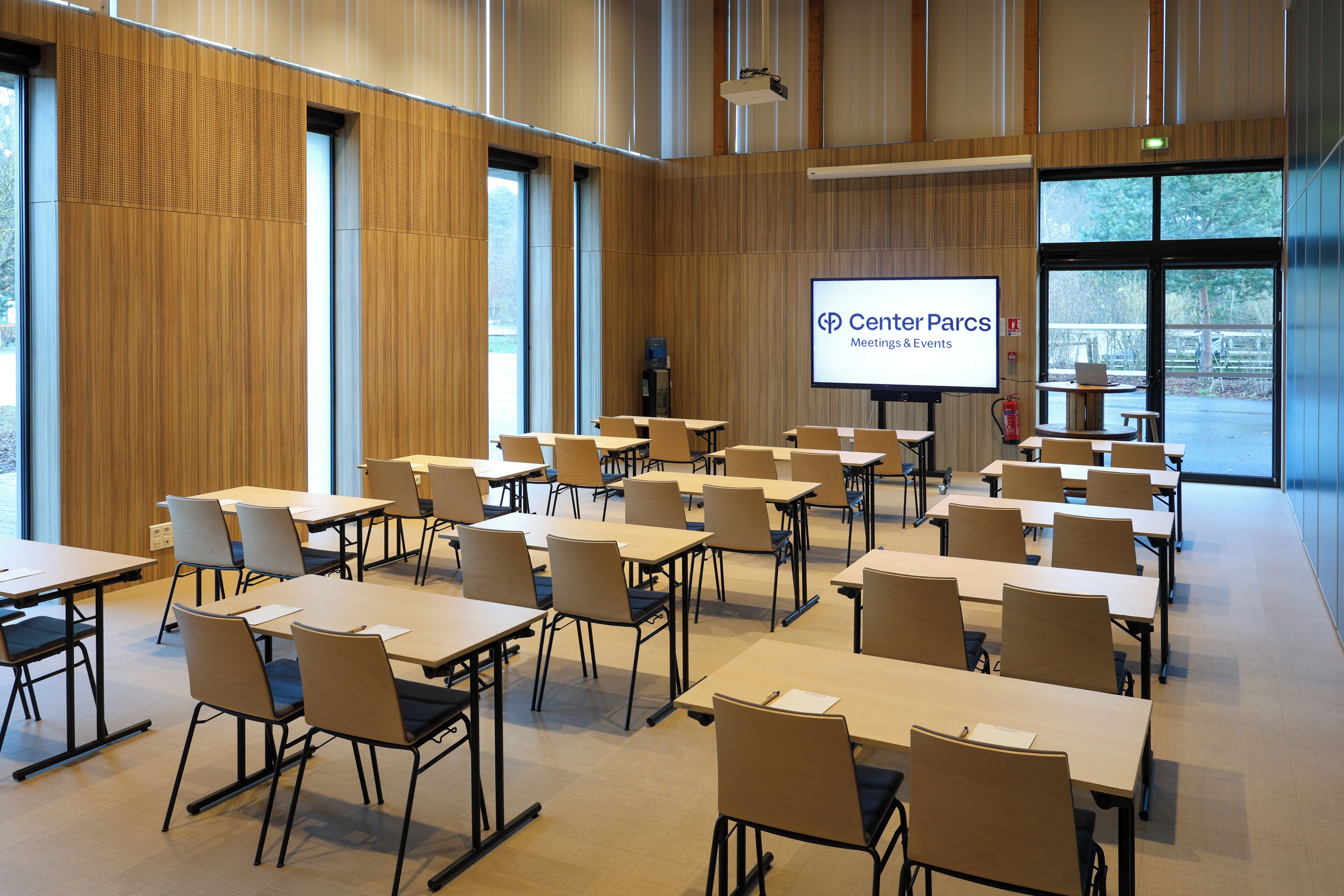 Ein Konferenzraum mit mehreren Tischen und Stühlen, einer Projektionsleinwand, auf der 'Center Parcs Meeting & Events' zu sehen ist, und großen Fenstern, die für natürliches Licht sorgen.