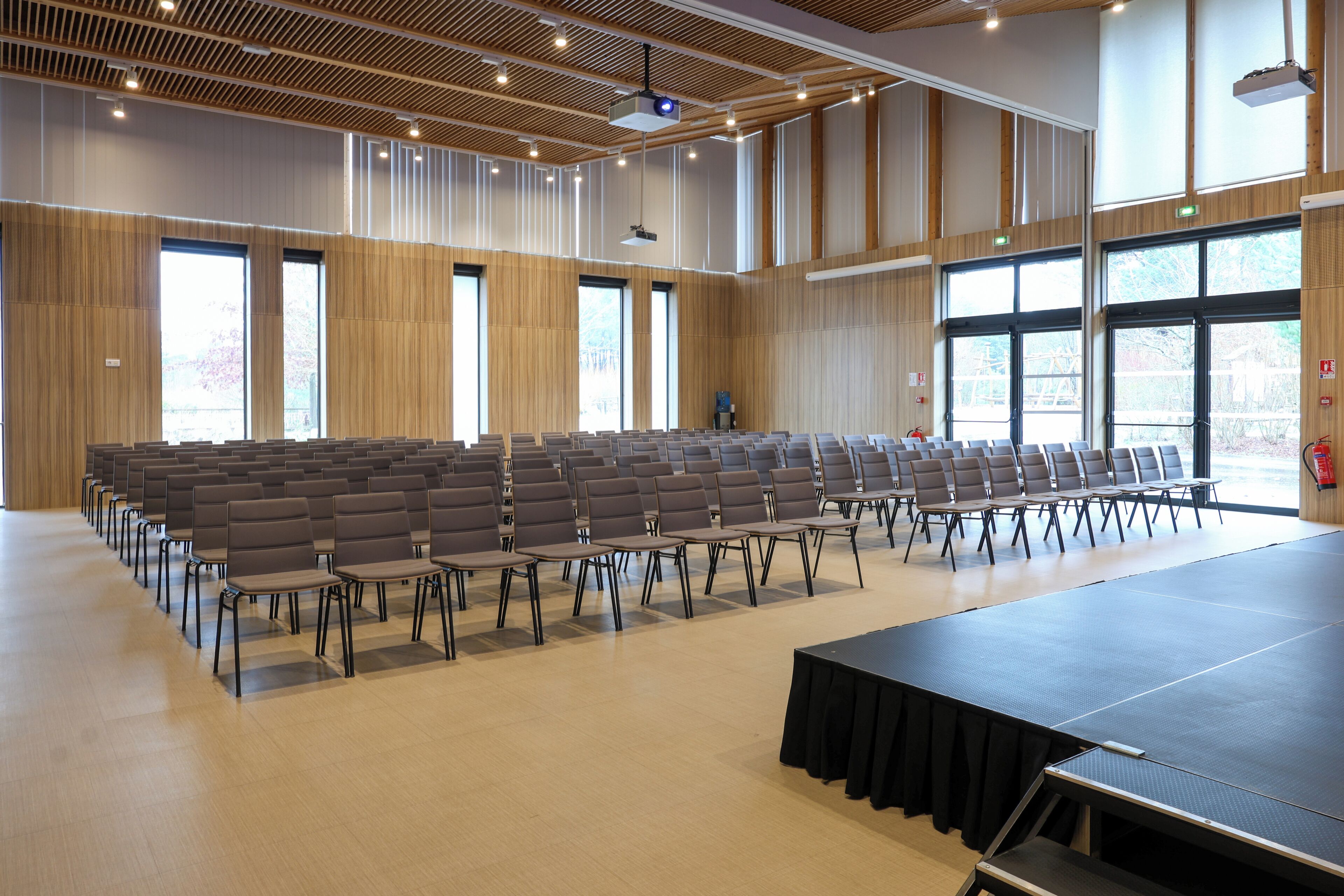 Uma sala de conferências com fileiras de cadeiras, um palco e grandes janelas.