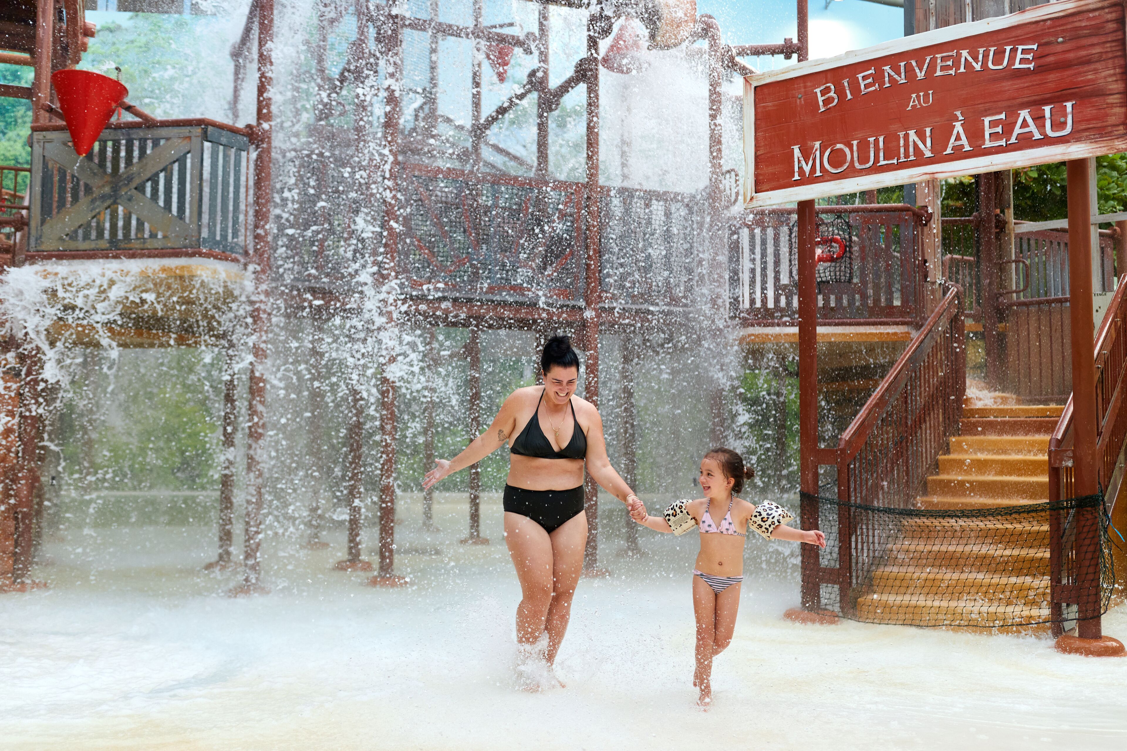 Une femme et un enfant se promènent dans un espace de jeux aquatiques, où un panneau indique : « Bienvenue au Moulin à Eau ».