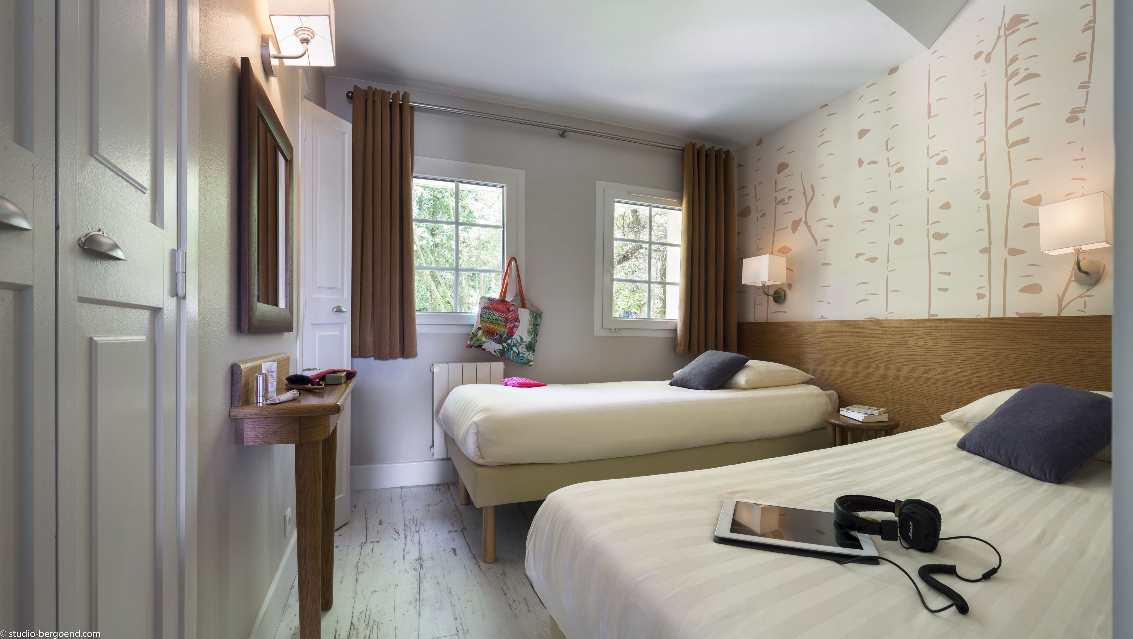 A hotel room with two beds, a bedside table with a lamp, a window with curtains, and a wall with a nature-themed wallpaper.