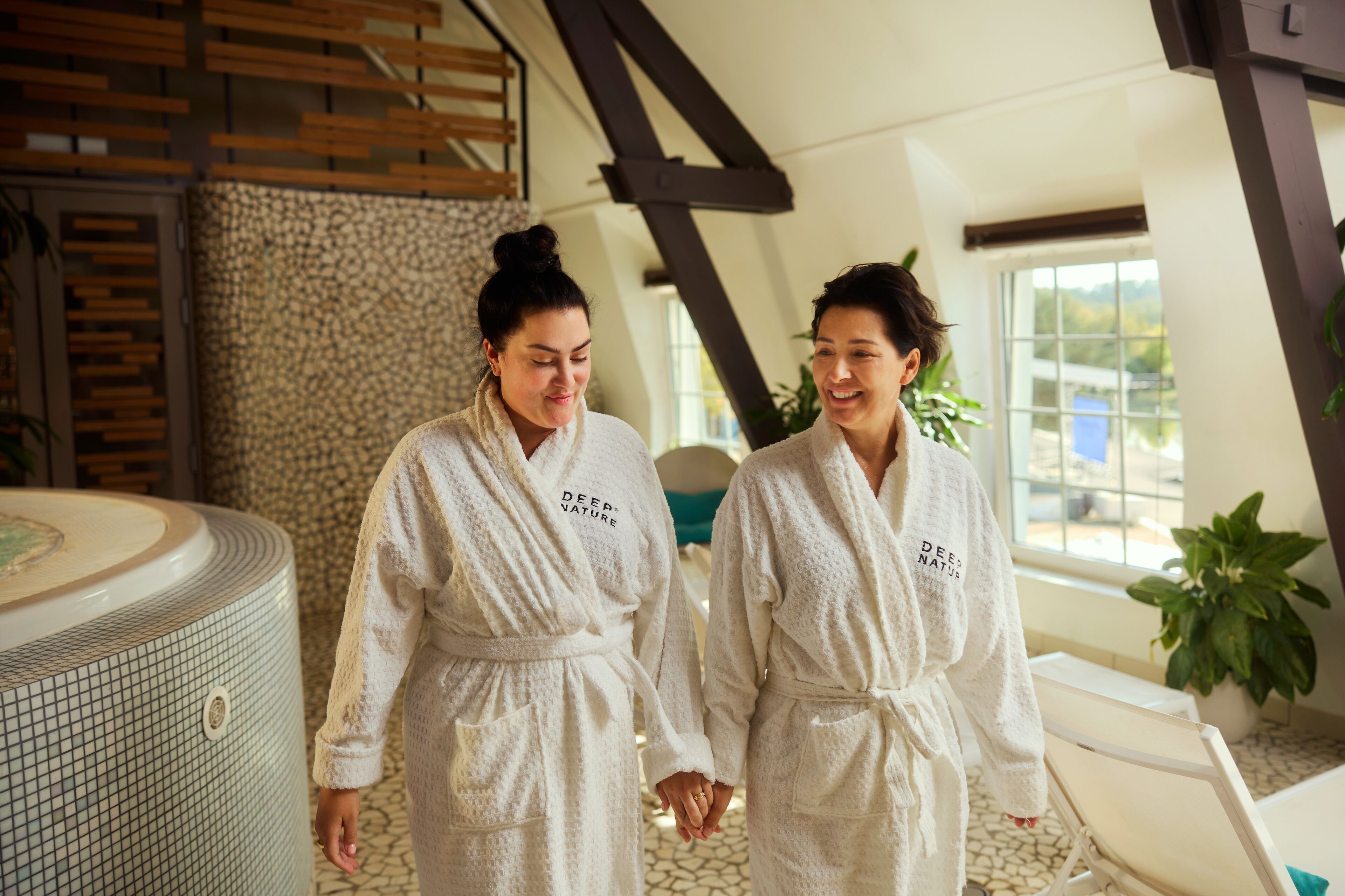 Two individuals in white robes with "DEEP NATURE" text, walking in a spa-like setting with a stone wall, a large bathtub, and a wooden ceiling structure.
